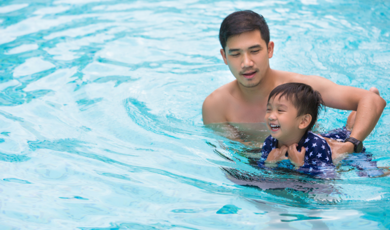Photo of father and preschool aged son in the water, both smiling as the father holds the child in the water.