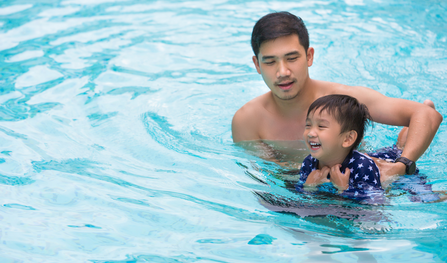 Photo of father and preschool aged son in the water, both smiling as the father holds the child in the water.